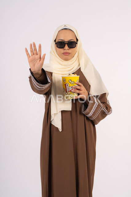 طلب التوقف عن فعل شيء ما،  تعابير الحزم والنهي، مشاهدة الأفلام داخل السينما، التسلية والترفيه بقضاء أوقات ممتعة، متابعة الفيلم المفضل ، الاستمتاع باكل البوب كورن وشرب المشروبات البارده ، بورتريه لامرأة عربية خليجية اماراتيه محجبة ترتدي عباءه وتحمل علبه فوشار ، خلفية بيضاء