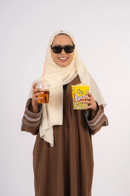 شرب المشروبات المنعشه ، اكل البوب كورن ، قضاء أوقات ممتعة ، الاستمتاع بمشاهدة الأفلام داخل السينما، مفهوم التسلية والترفيه ، متابعة الفيلم المفضل،بورتريه لامرأة عربية خليجية اماراتيه محجبة ترتدي عباءه وتحمل علبه فوشار ، خلفية بيضاء