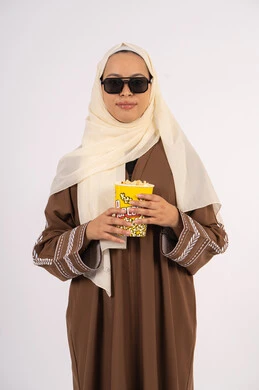 قضاء أوقات ممتعة ، اكل البوب كورن ، الاستمتاع بمشاهدة الأفلام داخل السينما، مفهوم التسلية والترفيه ، متابعة الفيلم المفضل،بورتريه لامرأة عربية خليجية اماراتيه محجبة ترتدي عباءه وتحمل علبه فوشار ، خلفية بيضاء