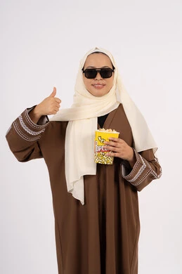 Enjoying watching movies in the cinema, expressions of approval and satisfaction, eating popcorn, entertainment and fun by spending a good time, following a favorite movie, portrait of a veiled Emirati Gulf Arab woman wearing an abaya and carrying a box of popcorn, white background