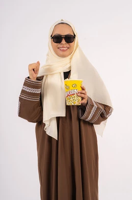 ايماءات الحماس والسرور ، رفع قبضة اليد بتعابير الفوز والانتصار ، بورتريه لامرأة عربية خليجية اماراتيه محجبة ترتدي عباءه ، تحمل كيس الفوشار، متابعة الفيلم المفضل، التسلية والترفيه بقضاء أوقات ممتعة، الاستمتاع بمشاهدة الأفلام داخل السينما ، خلفية بيضاء