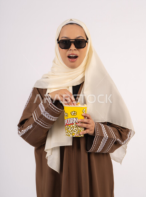 تعابير الصدمة والمفاجأة ، بورتريه لامرأة عربية خليجية اماراتيه محجبة ترتدي عباءه ، تحمل كيس الفوشار، متابعة الفيلم المفضل، التسلية والترفيه بقضاء أوقات ممتعة، الاستمتاع بمشاهدة الأفلام داخل السينما ، خلفية بيضاء