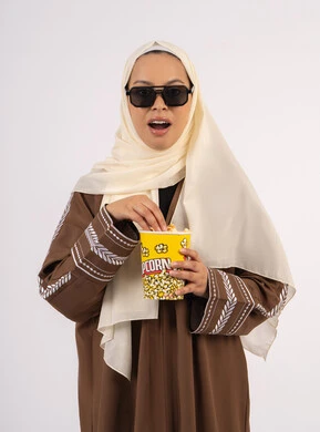 تعابير الصدمة والمفاجأة ، بورتريه لامرأة عربية خليجية اماراتيه محجبة ترتدي عباءه ، تحمل كيس الفوشار، متابعة الفيلم المفضل، التسلية والترفيه بقضاء أوقات ممتعة، الاستمتاع بمشاهدة الأفلام داخل السينما ، خلفية بيضاء