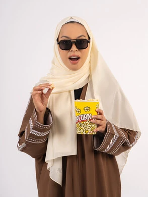 بورتريه لامرأة عربية خليجية اماراتيه محجبة ترتدي عباءه ، تحمل كيس الفوشار وترفع يدها بتعابير الموافقة ، ايماءات الجودة والامتياز، متابعة الفيلم المفضل، التسلية والترفيه بقضاء أوقات ممتعة، الاستمتاع بمشاهدة الأفلام داخل السينما ، خلفية بيضاء