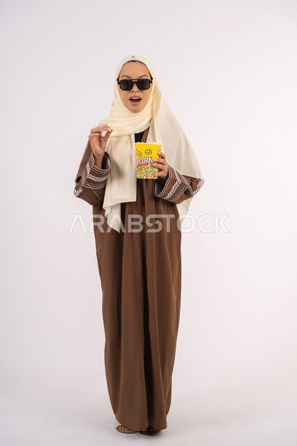 Gestures of quality and excellence, following a favorite movie, entertainment and fun, portrait of a veiled Emirati Gulf Arab woman wearing an abaya, holding a bag of popcorn and raising her hand in approval, enjoying watching movies inside the cinema, white background