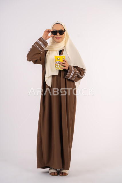 Watching your favorite movie, having fun and entertainment, looking through black glasses, portrait of a veiled Emirati Gulf Arab woman wearing an abaya, holding a bag of popcorn, enjoying watching movies inside the cinema, white background