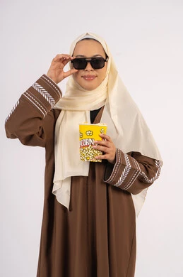 التسلية والترفيه بقضاء أوقات ممتعة، النظر من خلال نظاره سوداء ، بورتريه لامرأة عربية خليجية اماراتيه محجبة ترتدي عباءه ، تحمل كيس الفوشار ، الاستمتاع بمشاهدة الأفلام داخل السينما، الاستمتاع بتناول الفوشار ،متابعة الفيلم المفضل، خلفية بيضاء