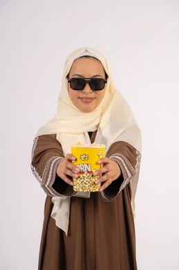 Enjoying watching movies in the cinema, wearing sunglasses, having fun and entertainment, enjoying popcorn, watching your favorite movie, portrait of a veiled Emirati Gulf Arab woman wearing an abaya, offering a bag of popcorn, white background