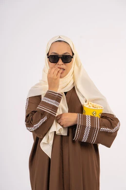 التسلية والترفيه بقضاء أوقات ممتعة، تعابير الاندماج والتفاعل ،الاستمتاع باكل حبات الفوشار ،متابعة الفيلم المفضل، الاستمتاع بمشاهدة الأفلام داخل السينما، بورتريه لامرأة عربية خليجية اماراتيه محجبة ترتدي عباءه ، تحمل كيس الفوشار ، خلفية بيضاء