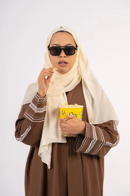 تعابير الاندماج والتفاعل ، متابعة الفيلم المفضل، الاستمتاع بمشاهدة الأفلام داخل السينما، بورتريه لامرأة عربية خليجية اماراتيه محجبة ترتدي عباءه ، وتأكل حبات الفوشار، التسلية والترفيه بقضاء أوقات ممتعة،  خلفية بيضاء