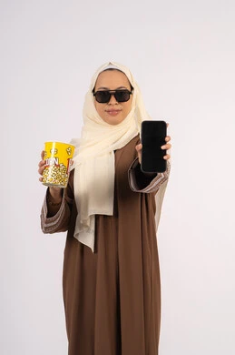 عرض شاشه سوداء فارغه ، الاستمتاع بمشاهدة الأفلام داخل السينما، بورتريه لامرأة عربية خليجية اماراتيه محجبة ترتدي عباءه وتحمل كيس فوشار والجوال ، التسلية والترفيه بقضاء أوقات ممتعة، ايماءات السعادة والسرور لمتابعة الفيلم المفضل، خلفية بيضاء