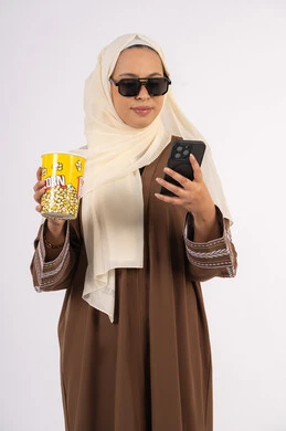 دمج التكنولوجيا الحديثه فى الحياه اليوميه ، الاستمتاع بمشاهدة الأفلام داخل السينما، بورتريه لامرأة عربية خليجية اماراتيه محجبة ترتدي عباءه وتحمل كيس فوشار والجوال ، التسلية والترفيه بقضاء أوقات ممتعة، ايماءات السعادة والسرور لمتابعة الفيلم المفضل، خلفية بيضاء