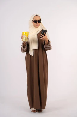 التواصل مع الاهل والاصدقاء ، الاستمتاع بمشاهدة الأفلام داخل السينما، بورتريه لامرأة عربية خليجية اماراتيه محجبة ترتدي عباءه وتحمل كيس فوشار والجوال ، التسلية والترفيه بقضاء أوقات ممتعة، ايماءات السعادة والسرور لمتابعة الفيلم المفضل، خلفية بيضاء