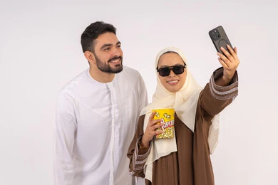 Using mobile phone to document memorable moments, enjoying eating popcorn, expressions of pleasure and happiness, portrait of a smiling Emirati Gulf Arab couple taking selfies, enjoying family entertainment activities, watching movies and series, having a good time at the cinema, white background