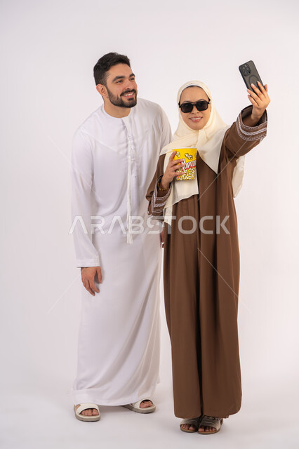 Integrating modern technology into daily life, enjoying popcorn, expressions of pleasure and happiness, portrait of a smiling Emirati Gulf Arab couple taking selfie, enjoying family entertainment activities, watching movies and series, having a good time at the cinema, white background
