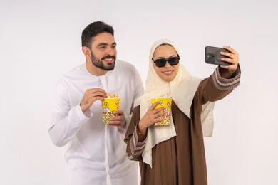 Love and affection between spouses, enjoying eating popcorn, expressions of pleasure and happiness, portrait of a smiling Emirati Gulf Arab couple looking at the camera, enjoying family entertainment activities, watching movies and series, having a good time at the cinema, white background