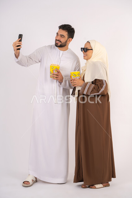 التقاط صور السيلفي ،بورتريه لزوجيين عربيين خليجيين اماراتيين مبتسميين ينظران الى الكاميرا، تعابير الاندماج والتفاعل،الاستمتاع بالأنشطة العائلية الترفيهيه، تناول الفوشار اثناء متابعة الأفلام والمسلسلات، قضاء وقت ممتع في السينما،خلفية بيضاء