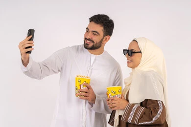 Documenting memorable moments via mobile, portrait of a smiling Emirati Gulf Arab couple looking at the camera, expressions of integration and interaction, enjoying family entertainment activities, eating popcorn while watching movies and series, having a good time at the cinema, white background