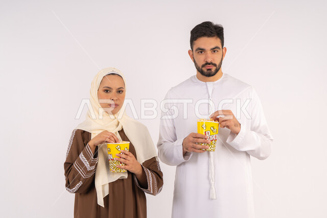 تعابير الاندماج والتفاعل،الاستمتاع بالأنشطة العائلية الترفيهيه، تناول الفوشار اثناء متابعة الأفلام والمسلسلات، قضاء وقت ممتع في السينما، بورتريه لزوجيين عربيين خليجيين اماراتيين مبتسميين ينظران الى الكاميرا خلفية بيضاء
