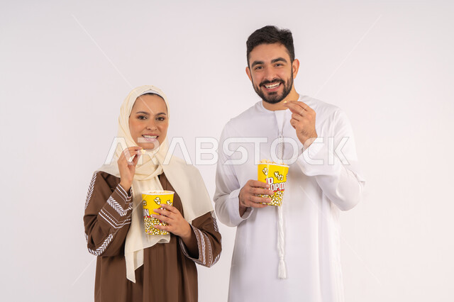 الاستمتاع بالأنشطة العائلية الترفيهيه، تناول الفوشار اثناء متابعة الأفلام والمسلسلات، قضاء وقت ممتع في السينما، بورتريه لزوجيين عربيين خليجيين اماراتيين مبتسميين ينظران الى الكاميرا بتعابير السرور والسعادة، خلفية بيضاء