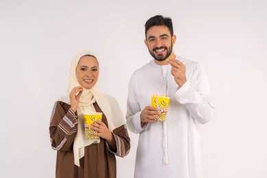 الاستمتاع بالأنشطة العائلية الترفيهيه، تناول الفوشار اثناء متابعة الأفلام والمسلسلات، قضاء وقت ممتع في السينما، بورتريه لزوجيين عربيين خليجيين اماراتيين مبتسميين ينظران الى الكاميرا بتعابير السرور والسعادة، خلفية بيضاء
