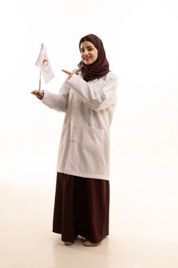 Contributing to humanitarian and social services, providing emergency medical first aid, the flag of the Saudi Red Crescent Authority, working in the medical field, a portrait of a Saudi Arabian Gulf female doctor wearing a white coat and hijab holding the Red Crescent flag in her hands and pointing to it with gestures of happiness, full-body, white background.