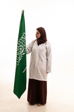 The Saudi flag is the banner of monotheism and Islam, commemorating the unification of the Kingdom of Saudi Arabia. A portrait of an Arab Saudi doctor wearing a coat and stethoscope looks at the Saudi flag, celebrating the National Day with pride and dignity, full-body, white background.