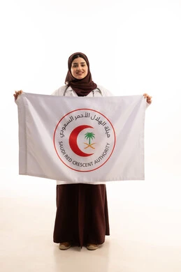 علم الهلال السعودي، مفهوم الطب والرعاية الصحية ، بورتريه لطبيبة عربية خليجية سعودية ترتدي العباءة والمئزر الأبيض تمسك بيديها علم عليه شعارالهلال الأحمر، النظر إلى الكاميرا بتعابير السعادة و السرور, الجسم بالطول الكامل، خلفية بيضاء