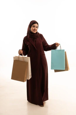 Shopping at commercial malls, looking at the camera with gestures of happiness and joy, excitement to take advantage of discounts on products, a portrait of a smiling Saudi Arabian woman wearing an abaya holding paper bags in her hand, taking advantage of seasonal offers and discounts and Black Friday, full-body shot, white background.