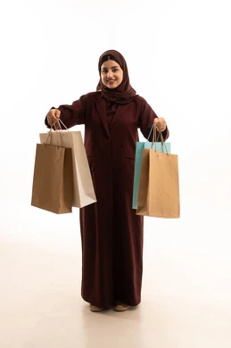 النظر الى الكاميرا بايماءات السعادة و السرور، الحماس للاستفادة من الخصومات على المنتجات، بورتريه لامرأة عربية سعودية مبتسمة ترتدي العباءة تحمل بيدها أكياس ورقية, استغلال العروض والتخفيضات الموسمية والجمعة البيضاء، التسوق من المولات التجارية، الجسم بالطول الكامل، خلفية بيضاء