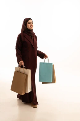 الحماس للاستفادة من الخصومات على المنتجات ، بورتريه لامرأة عربية سعودية مبتسمة ترتدي العباءة تحمل بيدها أكياس ورقية, استغلال العروض والتخفيضات الموسمية والجمعة البيضاء، التسوق من المولات التجارية، ايماءات السعاده و السرور، الجسم بالطول الكامل، خلفية بيضاء