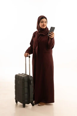قضاء اوقات ممتعة في الاجازة, بورتريه لامراة عربية خليجية ترتدي عباءة سوداء تحمل بيدها حقيبة سفر وهاتف محمول تنظر بايماءات السعادة و السرور, استخدام التقنيات الحديثة, الاستعداد للسياحة والسفر, الجسم بالطول الكامل، خلفية بيضاء