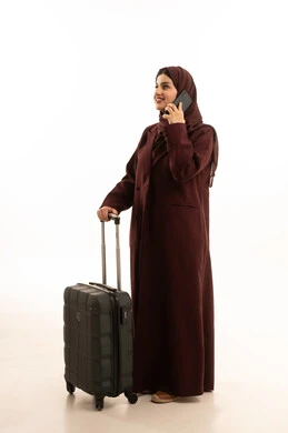 إجراء مكالمة هاتفية والدردشة مع الأصدقاء، الاستعداد للذهاب في رحلات ممتعة، استخدام الهاتف المحمول خلال وقت الانتظار، بورتريه لإمرأة عربية خليجية سعودية محجبة ترتدي العباءة تمسك بيديها حقيبة سفر، الجسم بالطول الكامل، خلفية بيضاء