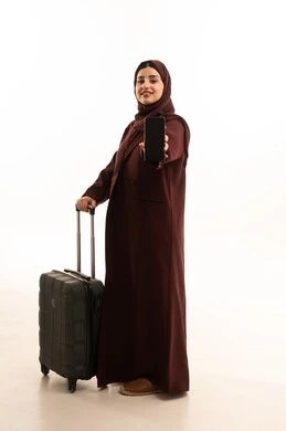 The use of modern and advanced devices, preparing for travel and enjoyable trips, a portrait of a Saudi Gulf Arab woman wearing a hijab and an abaya, holding a black travel bag and a mobile phone with a blank black screen, a full-body image, white background.