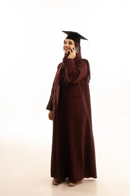 التواصل مع العائلة و الأصدقاء، استخدام جهاز تقني حديث, المناسبات والاحتفالات الجامعية, بورتريه لطالبة عربية سعودية خليجية محجبة ترتدي العباءة والقبعة تمسك بيدها الهاتف المحمول تقوم بإجراء مكالمة هاتفية, صورة الجسم بالكامل, خلفية بيضاء