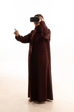 The development of technology and technology, the enjoyment and entertainment of watching through virtual reality glasses, a portrait of a Saudi Arabian Gulf woman wearing a hijab and abaya looking through 3D glasses (VR), the integration and simulation of the imaginary world, pointing to something, a full-body image, white background.
