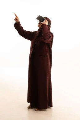 الاشارة الى شيء ما ، ايماءات التعجب و الاندهاش، تطور التقني والتكنولوجي, المتعة و التسلية بالمشاهدة عبر نظارة الواقع الافتراضي، بورتريه لامرأة عربية سعودية خليجية ترتدي الحجاب و العباءة تنظر من خلال نظارات ثلاثية الأبعاد (الفي ار), الاندماج ومحاكاة العالم الخيالي، صورة الجسم بالكامل، خلفية بيضاء
