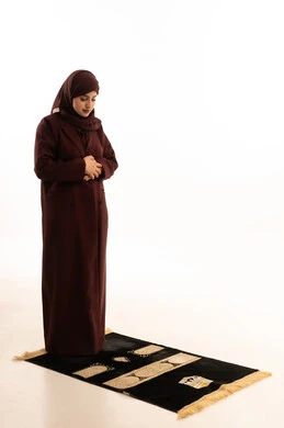 The concept of worship and drawing closer to God Almighty, striving in acts of obedience, supplications, and remembrances, maintaining the performance of the five daily prayers on time, a portrait of a Saudi Arabian Gulf woman wearing a hijab and an abaya standing on a prayer rug performing the obligatory prayer, full-body, white background.