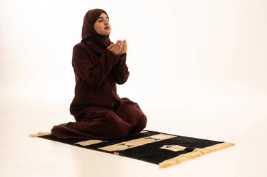 أداء الفروض على وقتها، تعابير تدل على التوسل والتضرع, التقرب الى الله من خلال الإجتهاد في العبادة والحفاظ على الصلوات، بورتريه لإمرأة عربية سعودية خليجية ترتدي الحجاب والعباءة البيضاء تجلس على سجادة الصلاة ترفع يديها وتدعو الله، خلفية بيضاء