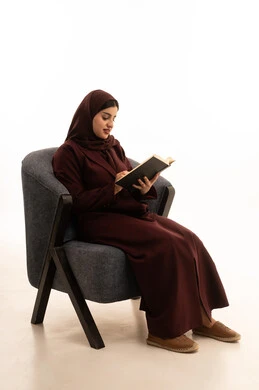 تعابير الاندماج و التركيز، حب المطالعة والقراءة, قضاء أوقات الفراغ بالتثقيف والتعلم, تنمية مهارات القراءة, الاهتمام بتصفح الكتب المفيدة,  بورتريه لسيدة عربية سعودية خليجية ترتدي الحجاب و العباءة تجلس على كرسي مريح تمسك بيدها الكتاب, خلفية بيضاء