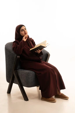 The concept of contemplation and thinking, the love of reading and literature, spending free time on education and learning, developing reading skills, the interest in browsing useful books, a portrait of an Arab Saudi Gulf woman wearing a hijab and abaya sitting on a comfortable chair holding a book in her hand, white background.