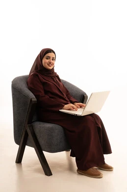 Looking at the camera with gestures of happiness and joy, completing remote work tasks, online shopping and orders, chatting and conversations and social networking, conducting a business meeting online, a portrait of a Saudi Arabian Gulf woman wearing a hijab and abaya sitting on a comfortable chair using a laptop, white background.