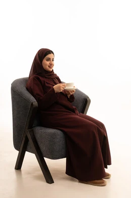 Spending free time sipping a favorite drink, gestures of pleasure and relaxation, enjoying hot beverages, consuming warm winter herbs, a portrait of a Saudi Arabian Gulf woman wearing a hijab and an abaya sitting on a comfortable chair drinking coffee, white background.