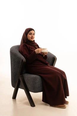 Gestures of enjoyment and relaxation, enjoying hot beverages, spending leisure time sipping a favorite drink, consuming warm winter herbs, a portrait of a Saudi Arabian Gulf woman wearing a hijab and an abaya sitting on a comfortable chair drinking coffee, white background.