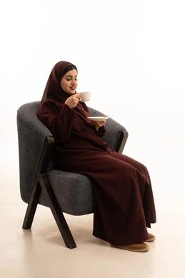 الاستمتاع بالمشروبات الساخنة، ايماءات المتعة والاسترخاء، قضاء وقت الفراغ باحتساء المشروب المفضل, تناول الأعشاب الشتوية الدافئة، بورتريه لامرأة عربية خليجية سعودية محجبة ترتدي العباءة تجلس على كرسي مريح بشرب القهوة، خلفية بيضاء