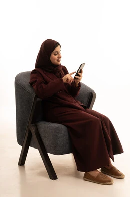 قضاء وقت ممتع عبر الجوال مع الأصحاب، تصفح مواقع التواصل الاجتماعي، بورتريه لامرأة عربية خليجية سعودية محجبة ترتدي العباءة تجلس على كرسي مريح تقوم بإرسال الرسائل النصية عبر الهاتف المحمول، خلفية بيضاء