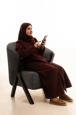 النظر الى شيء ما ، قضاء وقت ممتع عبر الجوال مع الأصحاب، تصفح مواقع التواصل الاجتماعي، بورتريه لامرأة عربية خليجية سعودية محجبة ترتدي العباءة تجلس على كرسي مريح تقوم بإرسال الرسائل النصية عبر الهاتف المحمول، خلفية بيضاء