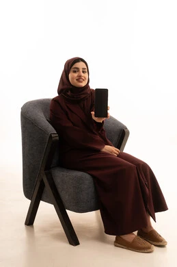 A display of a blank black screen, using a modern advanced technical device, a portrait of a smiling Saudi Arabian Gulf woman wearing a hijab and an abaya sitting on a comfortable chair holding a mobile phone in her hands looking at the camera with gestures of joy, white background.