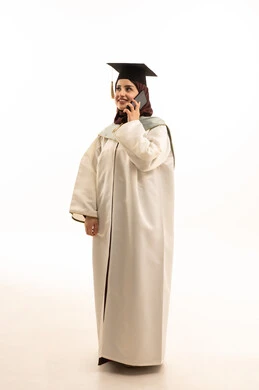 Using a modern technical device, university events and celebrations, a portrait of a Saudi Arabian Gulf female graduate wearing a white graduation gown and cap, holding a mobile phone making a phone call, full-body image, white background.