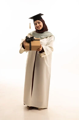 التعليم في المملكة العربية السعودية، الاحتفال بإنهاء الدراسة الجامعية، مفهوم الاجتهاد والتفوق، بورتريه لخريجة عربية سعودية خليجية محجبة ترتدي العباءة البيضاء وقبعة التخرج تحمل بيدها هدية تنظر الى الكاميرا بإيماءات الرضا والسرور ، الجسم بالطول الكامل، خلفية بيضاء